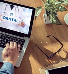 A person sitting at their laptop with the words “Dental” across the screen and glasses, a phone, notebook, pencils, and a cup of coffee nearby