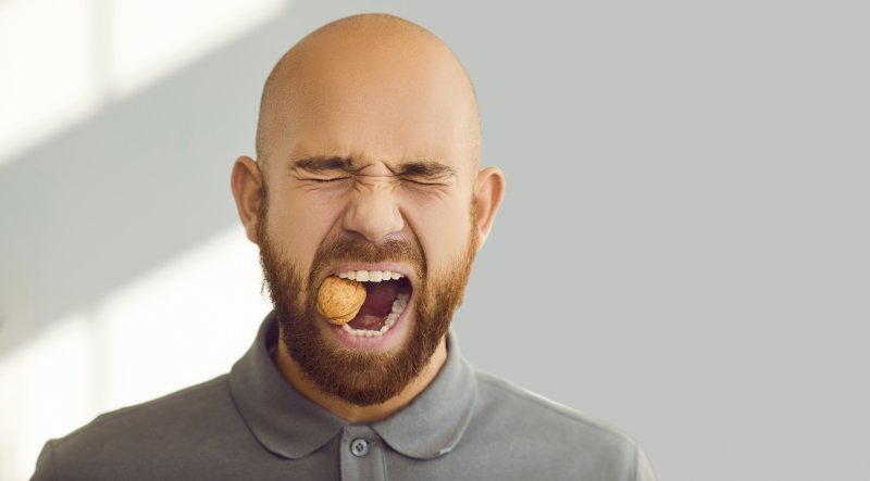 Man with strong healthy teeth trying to crack a walnut while standing on gray background.